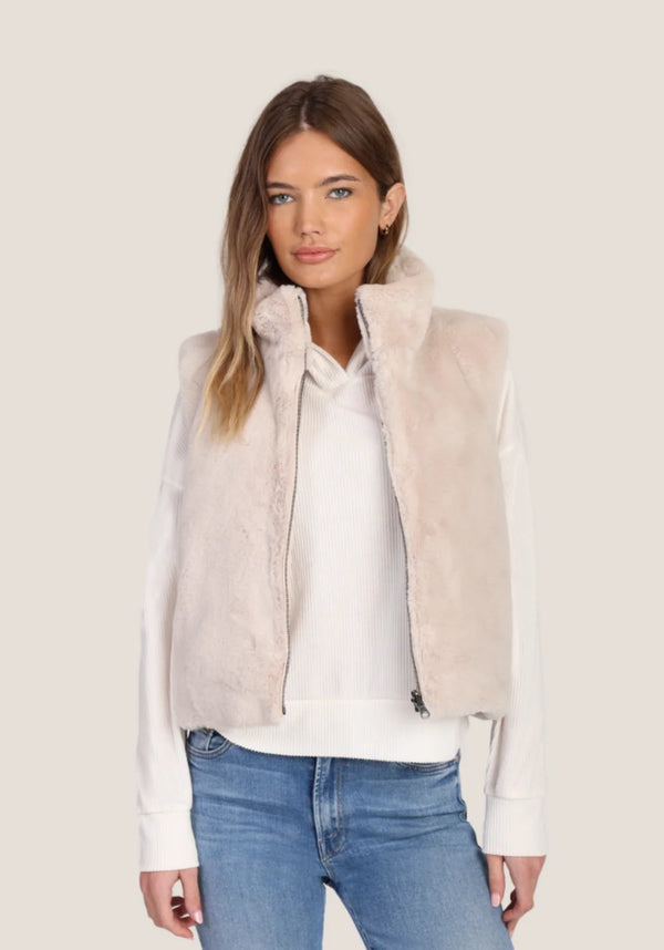 A woman modeling a light beige faux fur zip-up vest over a white long-sleeve top and blue jeans, set against a solid light beige background.