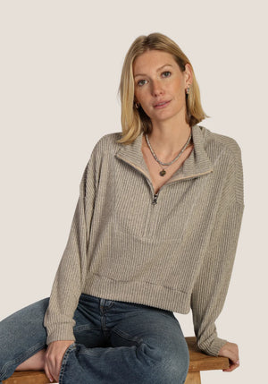 A woman with blonde hair sits on a wooden stool wearing a beige ribbed knit half-zip mock-neck sweater and blue jeans against a solid light beige background.