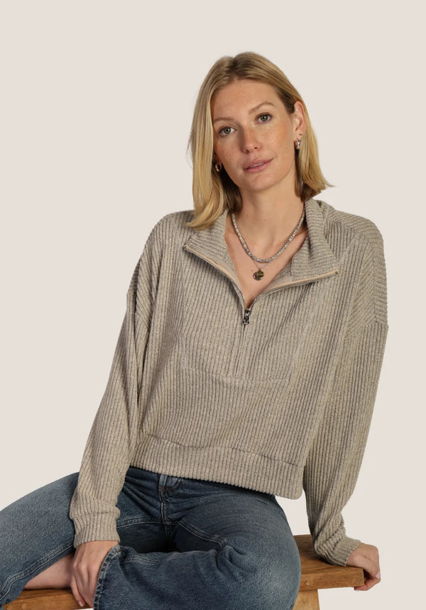 A woman with blonde hair sits on a wooden stool wearing a beige ribbed knit half-zip mock-neck sweater and blue jeans against a solid light beige background.