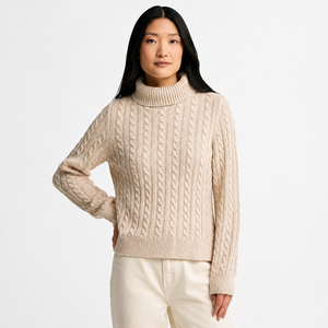 A woman wearing a cream-colored cable-knit turtleneck sweater and matching cream pants, photographed against a clean white studio background.