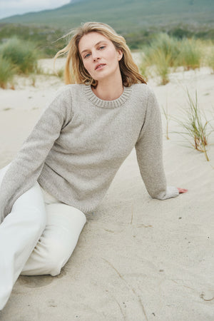 Woman sitting on a sandy beach wearing a light gray sweater and white pants.