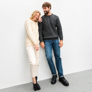 A man and a woman posing together against a white wall. The man wears a dark gray cable-knit crewneck sweater, blue jeans, and black boots.