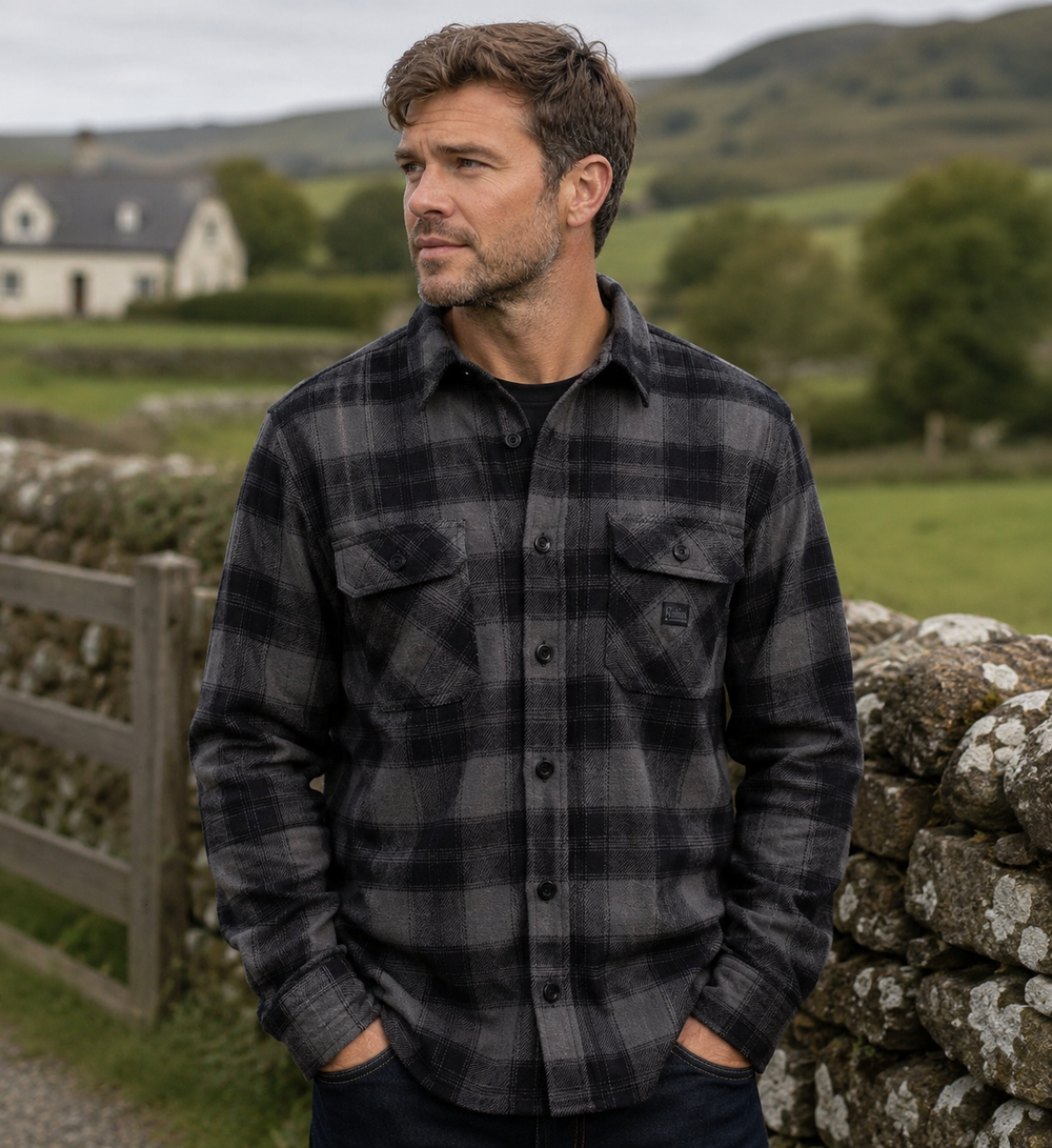 A man wearing a dark gray and black plaid flannel shirt stands outdoors in a rural setting with stone walls, green fields, and hills in the background.