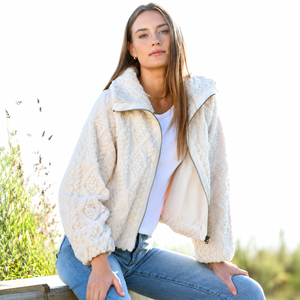 A woman wearing a cream-colored textured sherpa fleece jacket, a white t-shirt, and blue jeans, sitting outdoors against a bright, overexposed background.