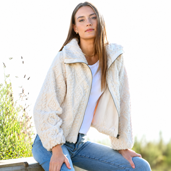 A woman wearing a cream-colored textured sherpa fleece jacket, a white t-shirt, and blue jeans, sitting outdoors against a bright, overexposed background.