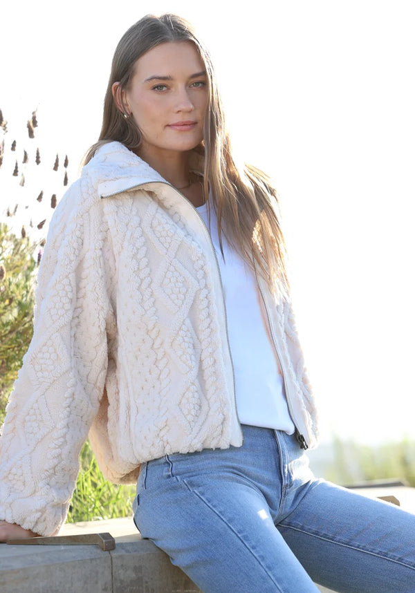 A woman wearing a cream cable-knit cropped jacket over a white top and blue jeans, sitting outdoors on a wooden ledge with bright sunlight and blurred greenery in the background.