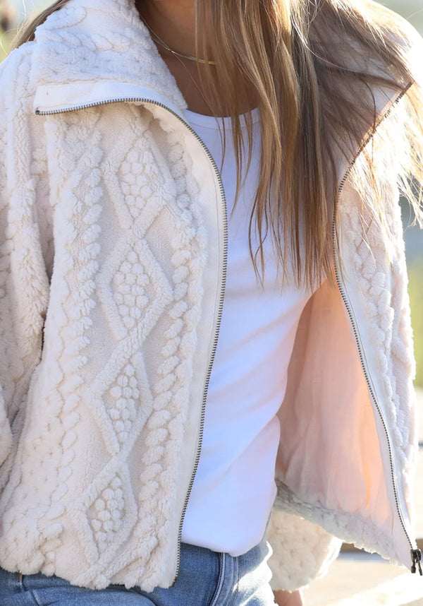 A close-up of a person wearing a cream-colored textured fleece zip-up jacket over a white t-shirt and blue jeans.