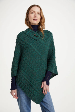A woman wearing a dark green cable-knit poncho sweater over a navy blue turtleneck and blue jeans, photographed against a plain white studio background.
