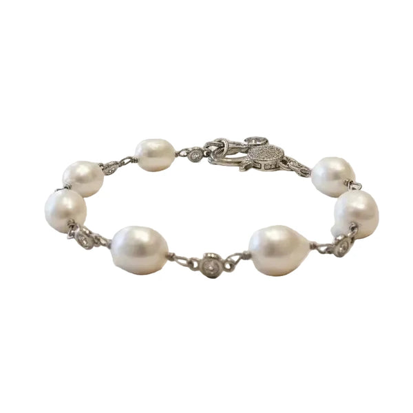 A silver chain bracelet featuring irregular white freshwater pearls and a round pavé clasp, displayed on a rustic white-washed wooden surface.