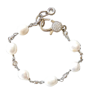 A silver chain bracelet featuring irregular white freshwater pearls and a round pavé clasp, displayed on a rustic white-washed wooden surface.
