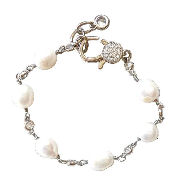 A silver chain bracelet featuring irregular white freshwater pearls and a round pavé clasp, displayed on a rustic white-washed wooden surface.