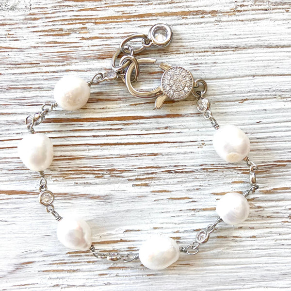A silver chain bracelet featuring irregular white freshwater pearls and a round pavé clasp, displayed on a rustic white-washed wooden surface.