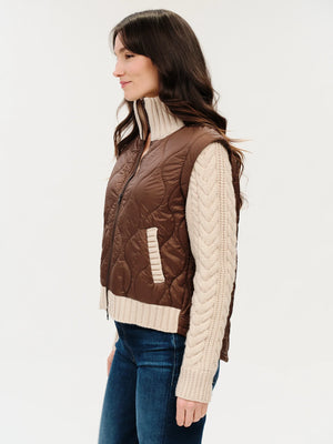 A woman shown in side profile wearing a brown quilted sleeveless vest over a cream cable-knit turtleneck sweater and blue jeans, against a plain light background.