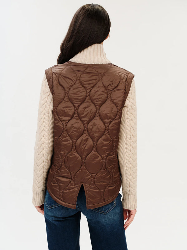 A woman shown from the back wearing a brown quilted puffer vest over a cream cable-knit turtleneck sweater and blue jeans, against a plain white studio background.