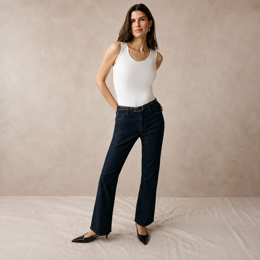 A woman models a pair of dark blue straight-leg jeans with a slim black belt, paired with a white tank top and black pointed-toe flats, standing against a textured beige studio backdrop.