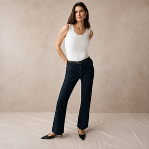 A woman models a pair of dark blue straight-leg jeans with a slim black belt, paired with a white tank top and black pointed-toe flats, standing against a textured beige studio backdrop.