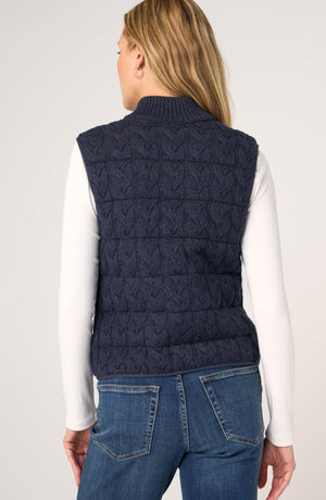 A woman shown from the back wearing a navy blue cable-knit sleeveless vest over a white long-sleeve top and blue jeans, against a light gray studio background.