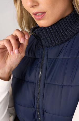 Close-up of a woman wearing a navy blue puffer vest with a ribbed knit high collar and zipper, with her hand adjusting the zipper.