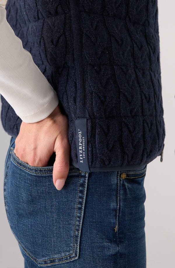 Close-up of a person wearing a dark navy cable-knit zip-front vest over a white long-sleeve top and blue jeans, with a small vertical Liverpool label visible at the side seam.