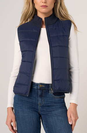 A woman wearing a navy blue sleeveless puffer vest over a white long-sleeve top and blue jeans, shown from the head down against a light gray studio background.