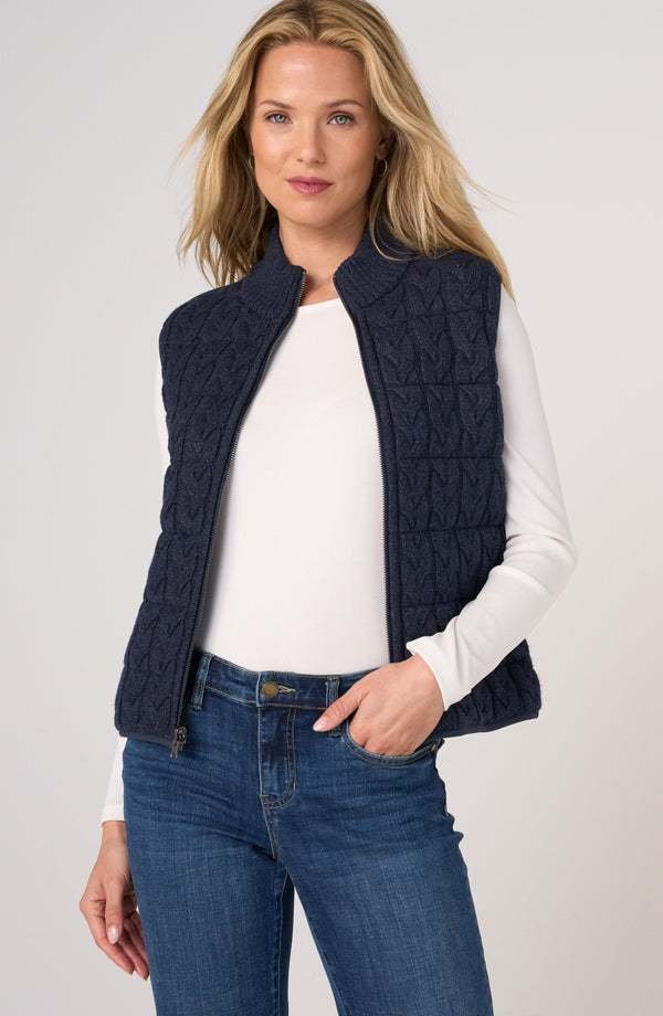 A woman wearing a navy blue quilted sleeveless vest over a white long-sleeve top and blue jeans, standing against a light gray studio background.