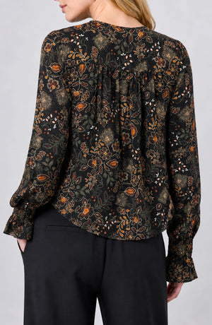 A woman wearing a black long-sleeve blouse with an intricate brown, orange, and gold floral pattern, shown from the back. The blouse features a gathered back neckline and ruffled cuffs, paired with black trousers against a light gray studio background.