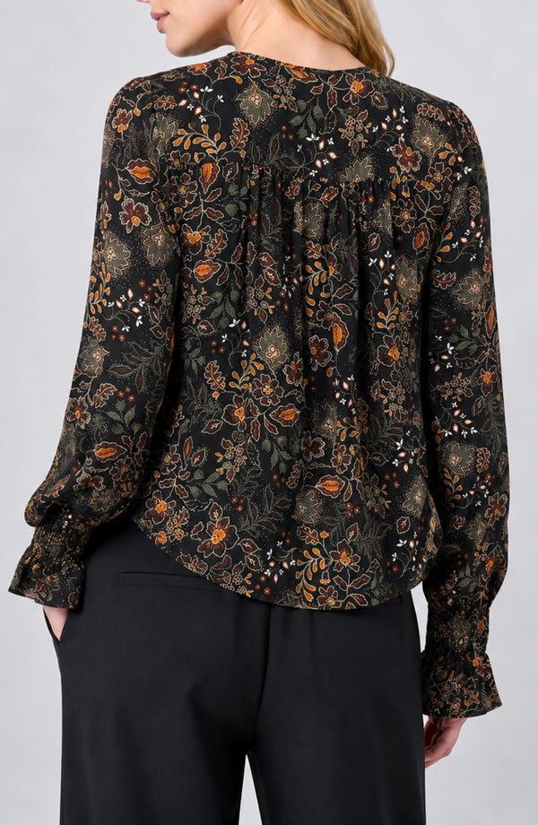 A woman wearing a black long-sleeve blouse with an intricate brown, orange, and gold floral pattern, shown from the back. The blouse features a gathered back neckline and ruffled cuffs, paired with black trousers against a light gray studio background.