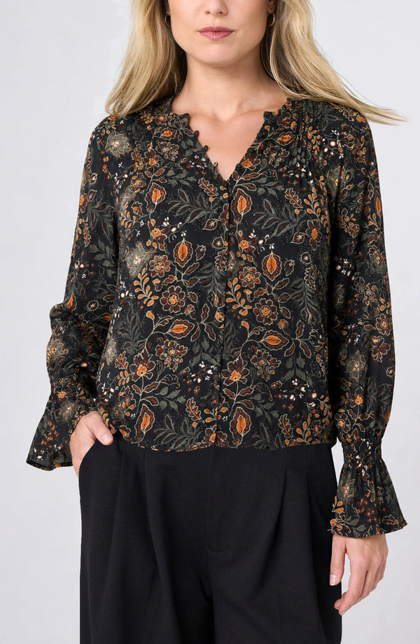 A woman wearing a black long-sleeve blouse with an intricate orange, brown, and green floral pattern, paired with black trousers, against a light gray studio background.