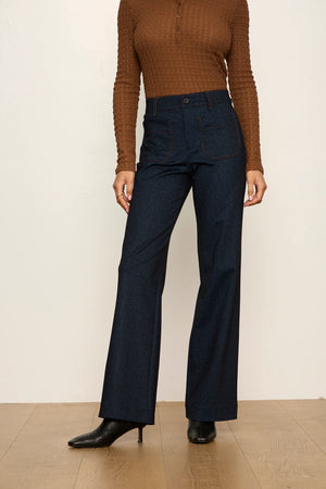 A model wearing dark blue high-waisted wide-leg jeans, a brown ribbed long-sleeve top, and black pointed-toe heels, standing against a plain off-white wall and light wood floor.