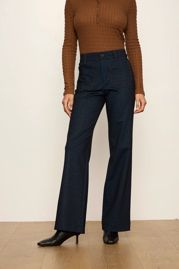 A model wearing dark blue high-waisted wide-leg jeans, a brown ribbed long-sleeve top, and black pointed-toe heels, standing against a plain off-white wall and light wood floor.