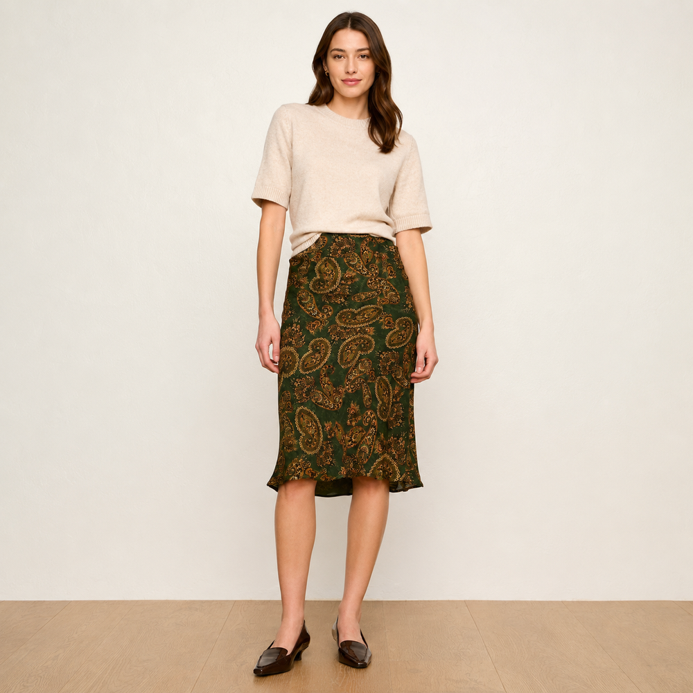 A woman stands against a plain off-white wall wearing a light beige short-sleeve knit top and a dark green midi skirt with an ornate gold paisley pattern. She is wearing dark brown loafers and standing on a light wood floor.