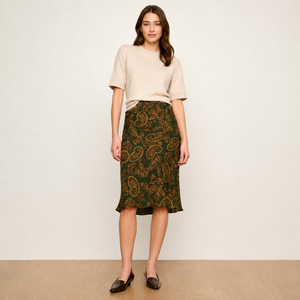 A woman stands against a plain off-white wall wearing a light beige short-sleeve knit top and a dark green midi skirt with an ornate gold paisley pattern. She is wearing dark brown loafers and standing on a light wood floor.