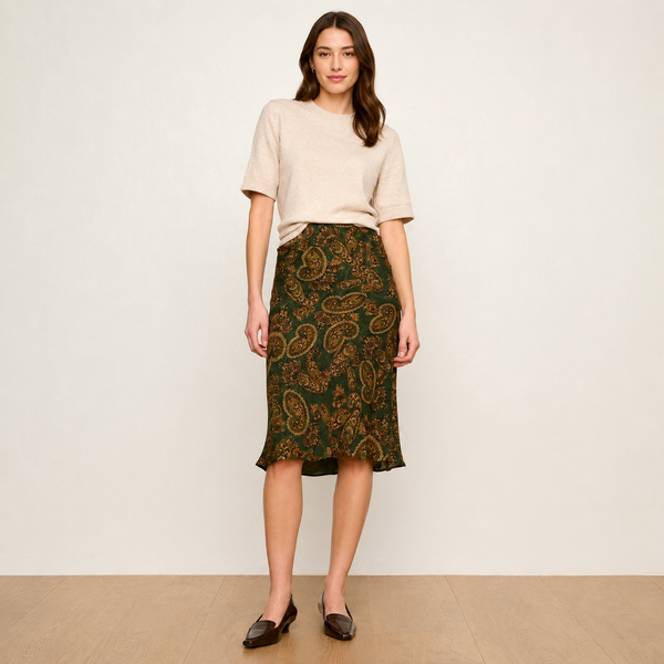 A woman stands against a plain off-white wall wearing a light beige short-sleeve knit top and a dark green midi skirt with an ornate gold paisley pattern. She is wearing dark brown loafers and standing on a light wood floor.