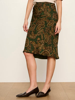 A model wearing a knee-length green and gold paisley skirt with black pointed-toe flats, standing against a plain beige wall.