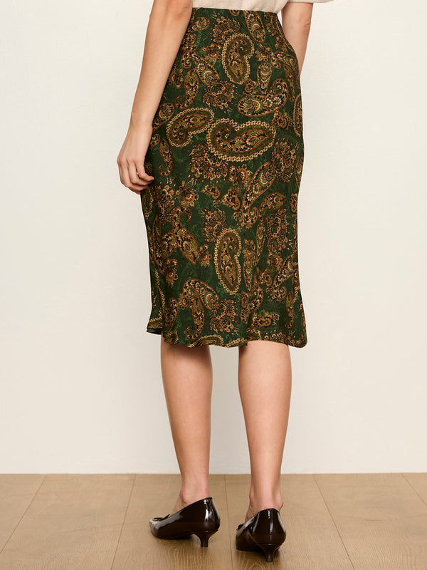 A model is shown from the back wearing a knee-length green skirt with a gold paisley pattern, paired with black heels, against a plain cream wall.