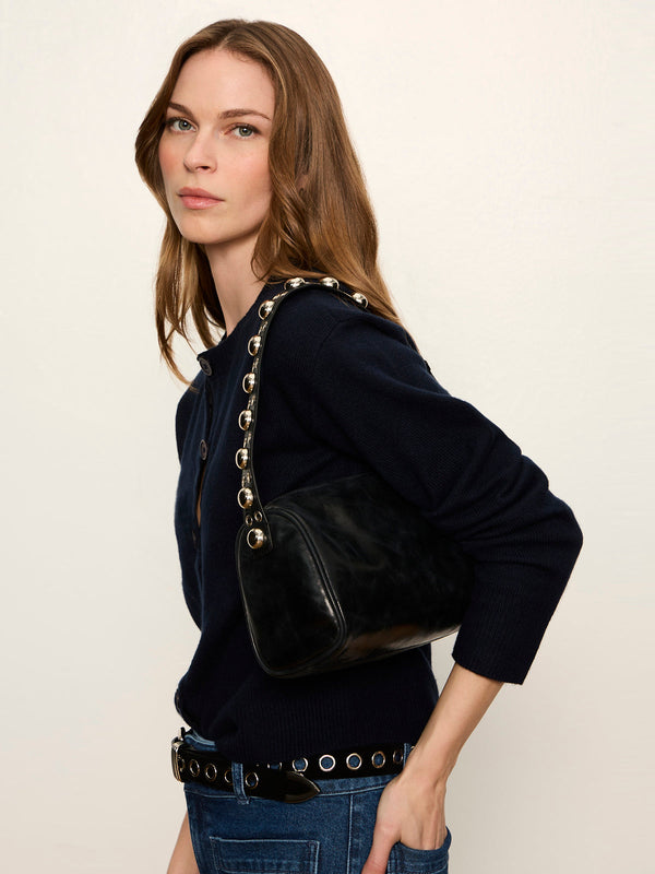 A woman wearing a navy blue knit top and jeans carries a black shoulder bag with a distinctive gold-tone chain strap against a plain off-white background.