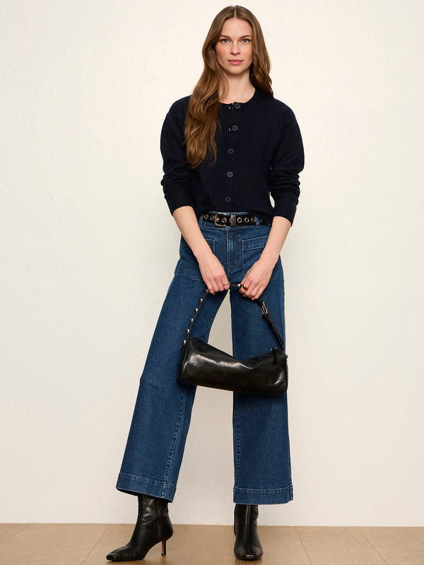 A woman modeling a dark navy cardigan, blue wide-leg jeans, black ankle boots, and holding a black leather shoulder bag against a plain beige background.