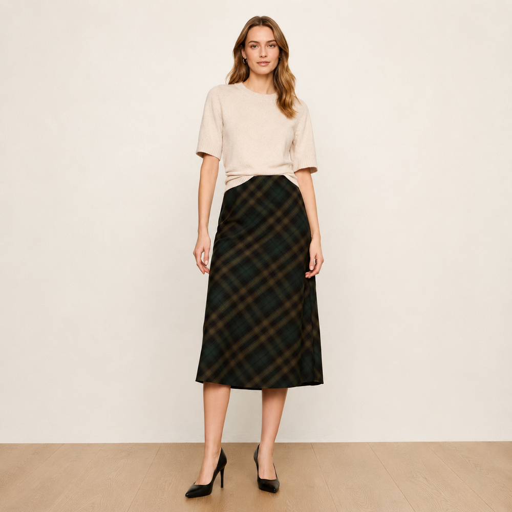 A woman stands in a studio wearing a light beige short-sleeve knit top and a dark green plaid midi skirt with black high heels.