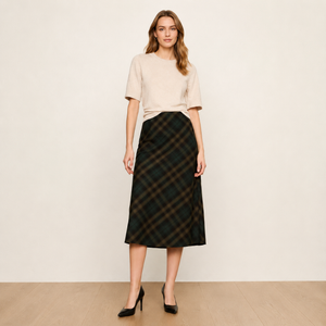 A woman stands in a studio wearing a light beige short-sleeve knit top and a dark green plaid midi skirt with black high heels.