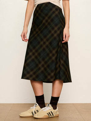 A person wearing a dark plaid midi skirt with a beige top, black socks, and beige sneakers, standing against a plain off-white wall.