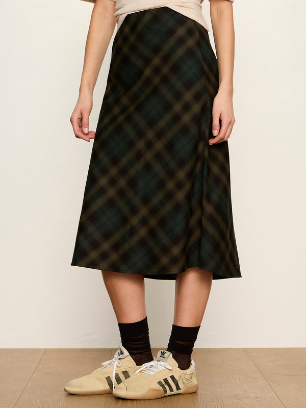 A person wearing a dark plaid midi skirt with a beige top, black socks, and beige sneakers, standing against a plain off-white wall.