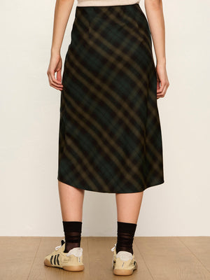 A model from the waist down wearing a dark green and black plaid midi skirt, paired with black socks and beige sneakers, standing against a plain off-white wall.