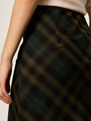 Close-up of a person wearing a dark plaid skirt with green, black, and brown tones, paired with a light beige top against a plain light background.