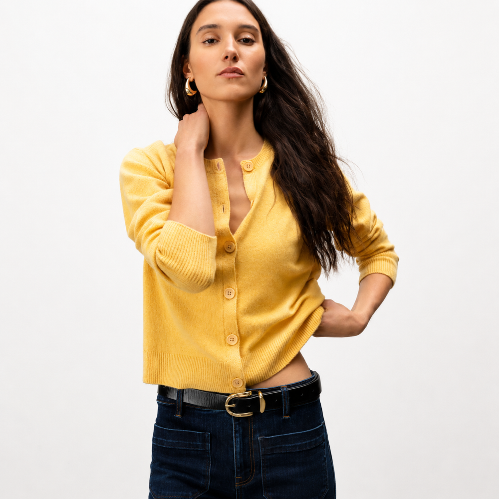 A woman modeling a cropped yellow ribbed cardigan with buttons, paired with dark blue jeans and a black belt, against a plain white studio background.
