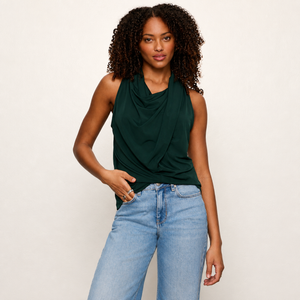 A woman with curly dark hair wearing a dark green sleeveless draped top and light blue jeans, standing against a plain off-white studio background.
