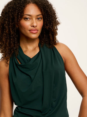 A woman with curly dark hair wearing a dark green sleeveless draped top, shown from the chest up against a plain light background.