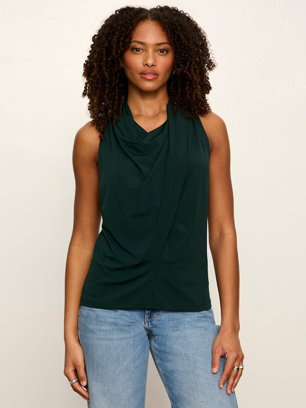 A woman with curly dark hair wearing a dark green sleeveless cowl-neck top and light blue jeans, standing against a plain off-white studio background.