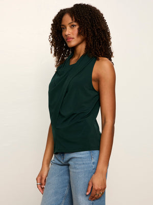 A woman with curly dark hair wearing a dark green sleeveless draped top and light blue jeans, standing against a plain off-white studio background.
