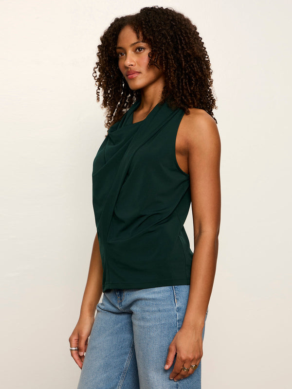 A woman with curly dark hair wearing a dark green sleeveless draped top and light blue jeans, standing against a plain off-white studio background.