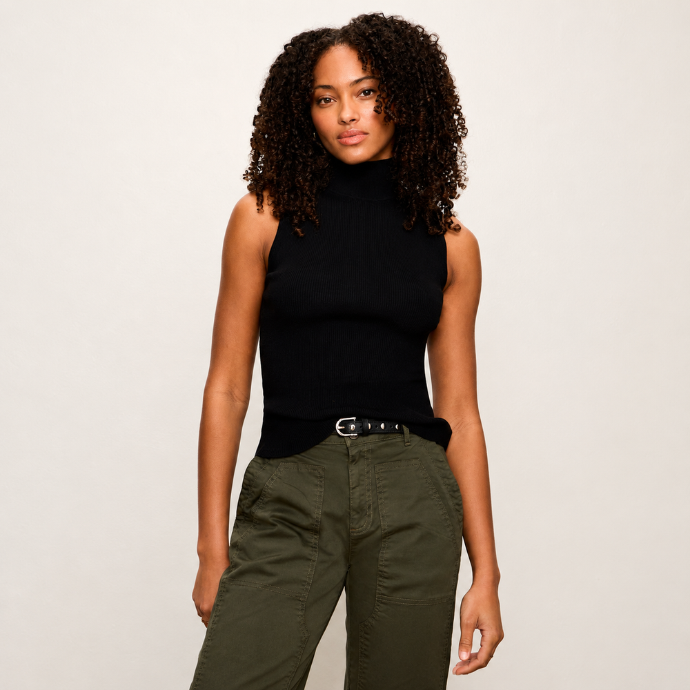 A woman with curly dark hair wearing a black sleeveless mock neck top and olive green cargo pants, standing against a light gray studio background.